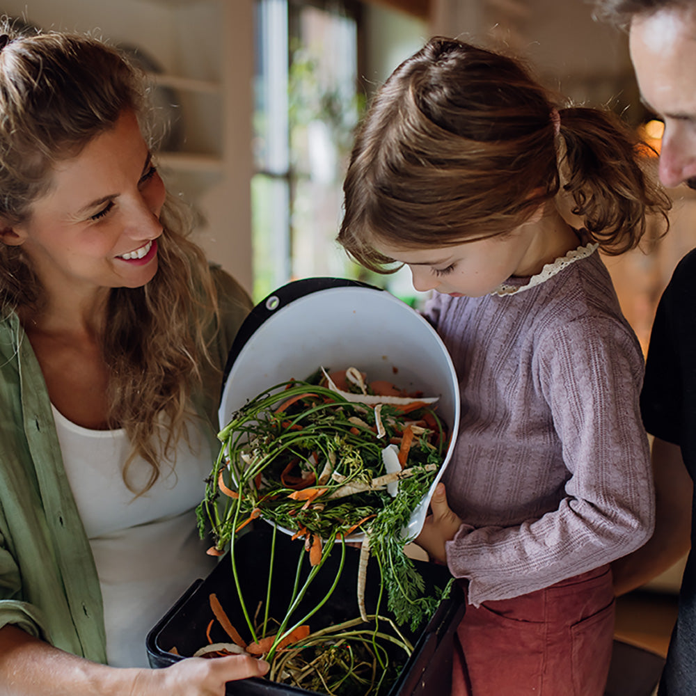 A woman and a child are seen emptying food scraps, including carrot tops and vegetable remnants, from a small kitchen compost bin. The child holds the bin while the woman assists, symbolizing family participation in sustainable practices. This image conveys the importance of composting food waste and teaching environmental responsibility to future generations. Compostable waste bags can be a key tool in supporting kitchen composting efforts, keeping food scraps contained and ready for proper disposal.